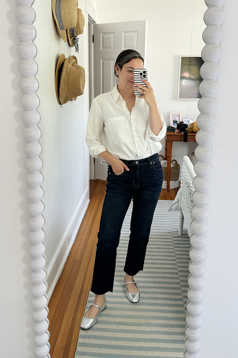 A mirror selfie of Carly wearing dark denim jeans, metallic silver mary janes, and an oversized white button down.