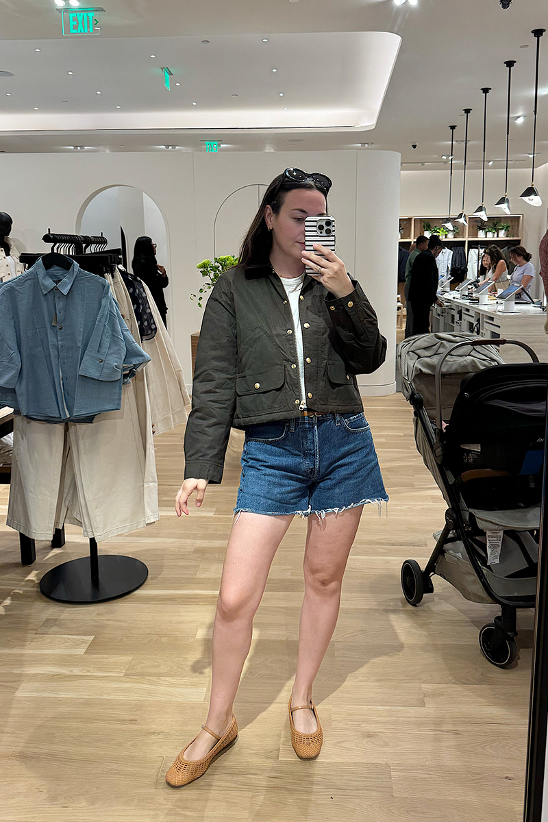 A mirror selfie of Carly wearing an olive green short barn jacket and denim shorts and ballet flats. 