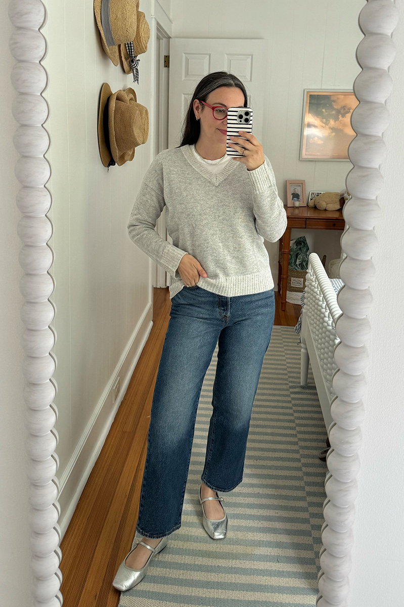 Carly Riordan taking a mirror selfie wearing a grey sweater and jeans and metallic ballet flats. 