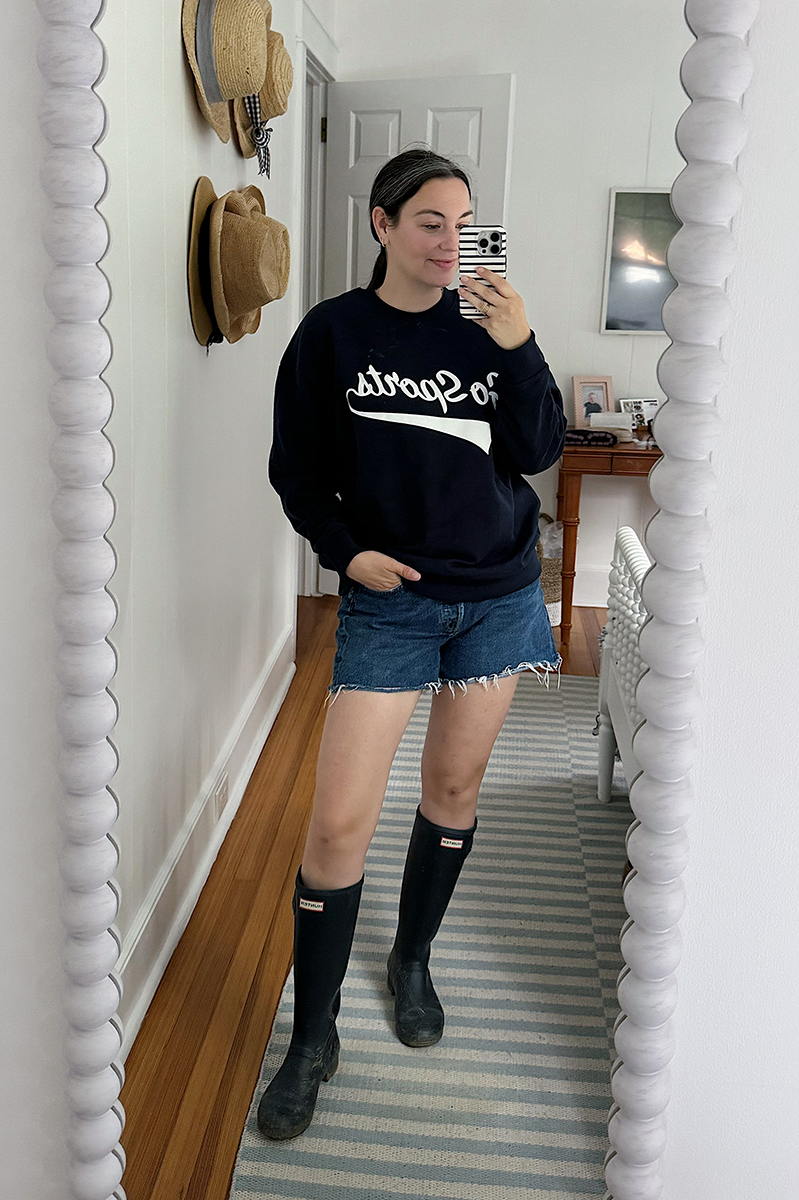 Carly taking a mirror selfie wearing denim shorts, tall rainboots, and a navy sweatshirt.