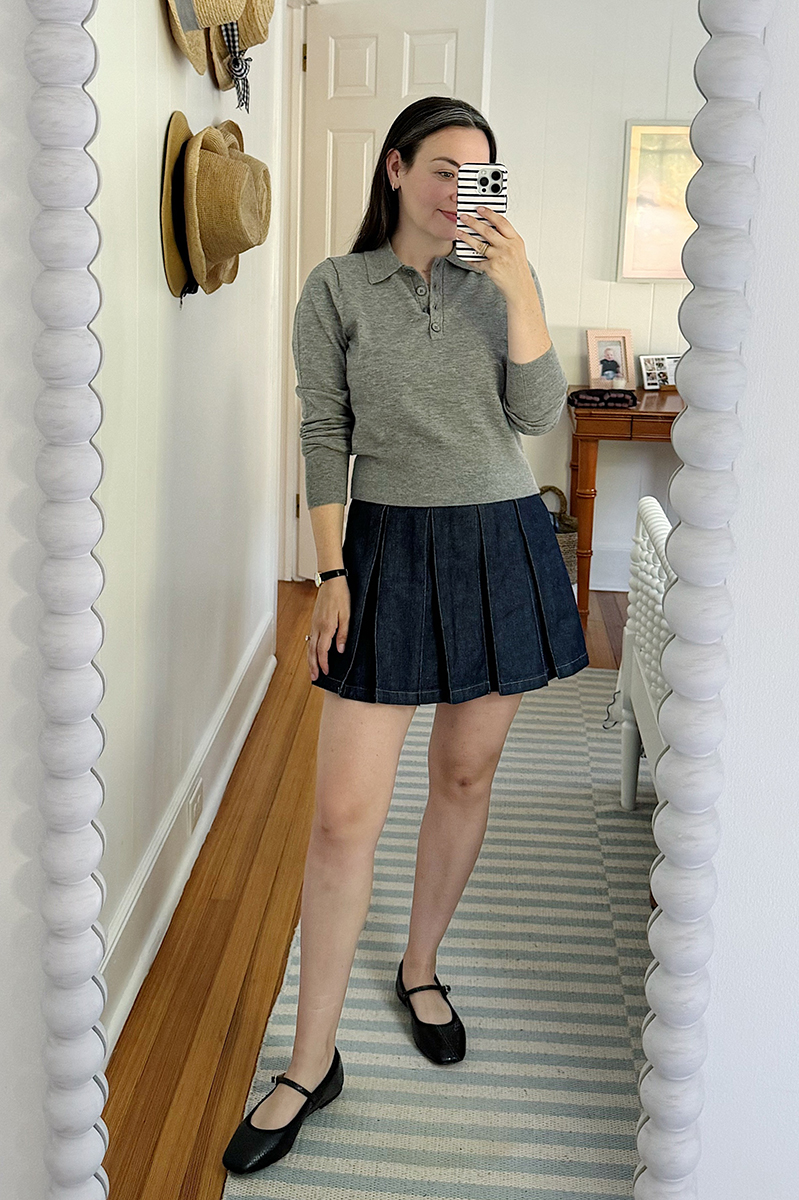 Carly taking a mirror selfie wearing a denim pleated skort and grey polo sweater. 