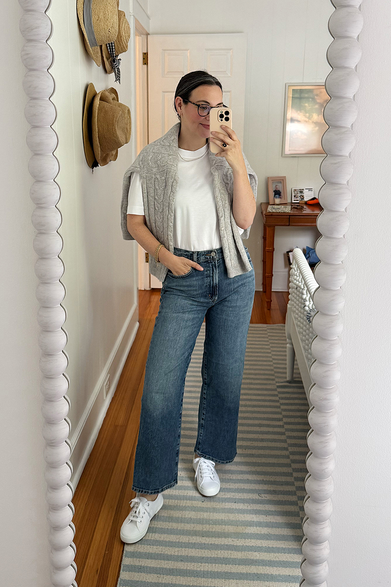 Carly in a mirror selfie wearing a white tee, grey sweater, and wide legged jeans. 