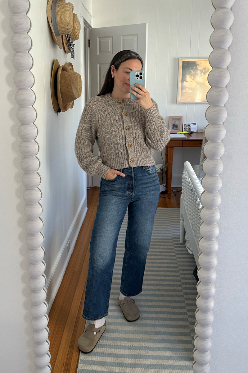 Carly Riordan taking a mirror selfie wearing a cardigan, jeans, and Boston clogs.