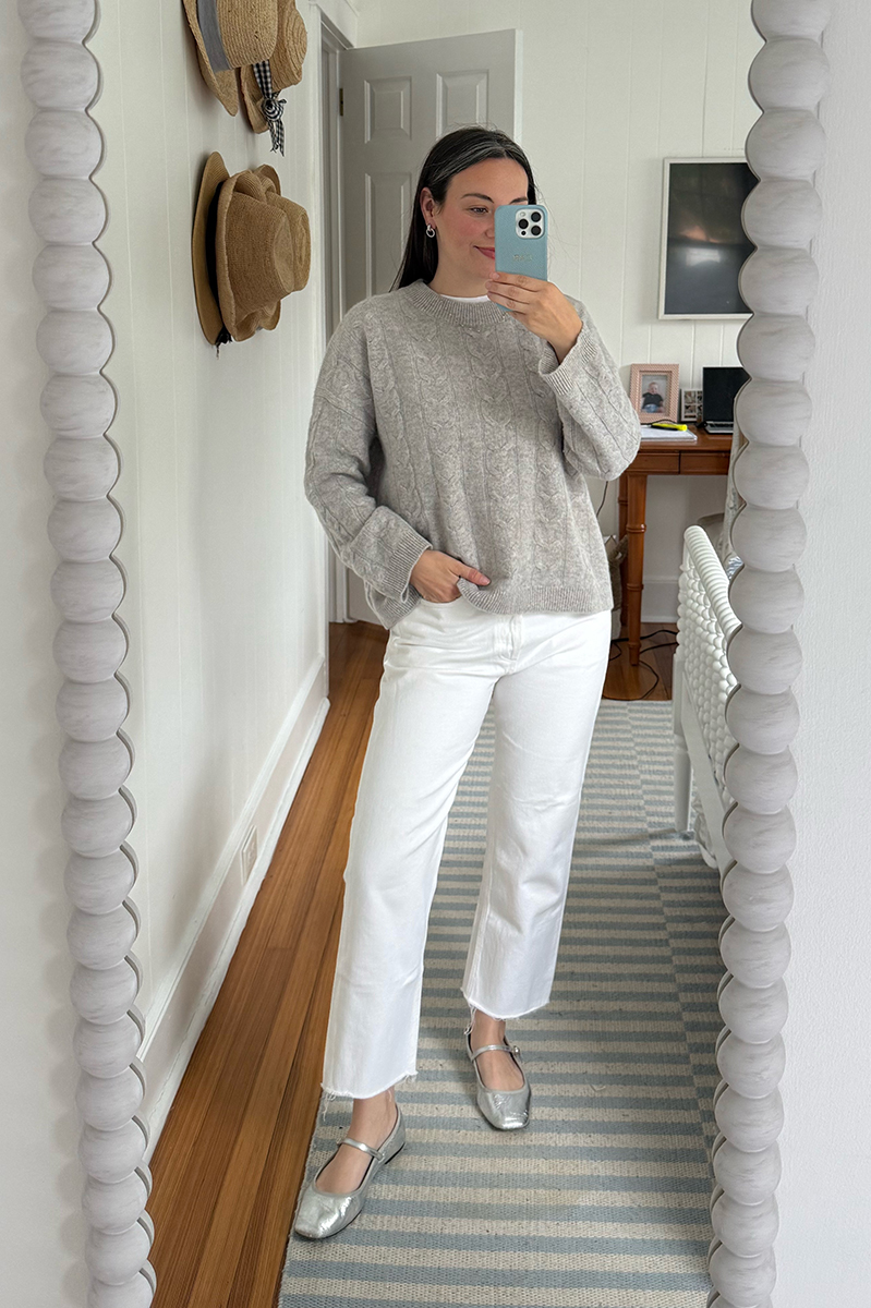 Carly Riordan taking a mirror selfie wearing a grey sweater, white jeans, and silver mary janes.