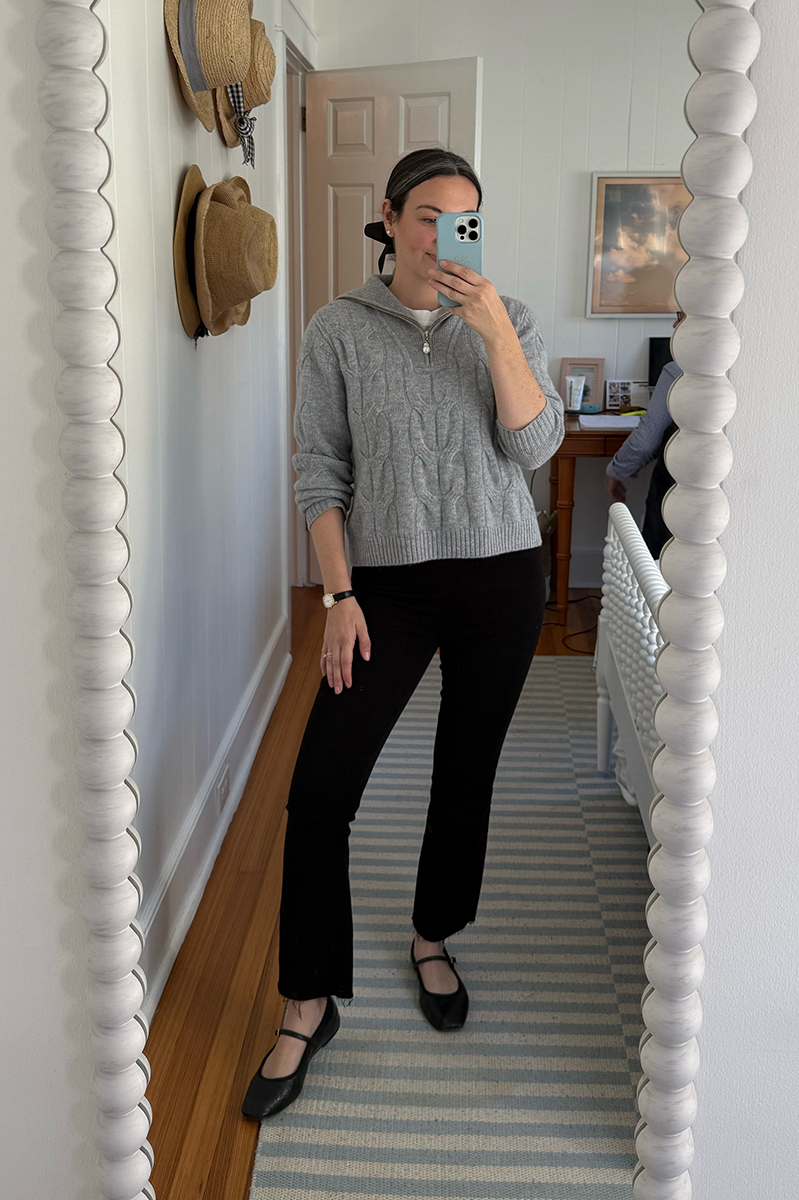 Carly Riordan taking a mirror selfie wearing a grey pullover, black jeans, and mary janes.