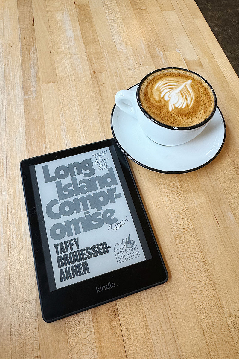 A kindle with Long Island Compromise loaded and a cappuccino on a table.