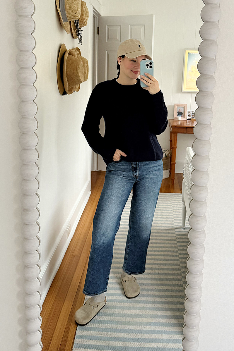 Carly Riordan taking a mirror selfie wearing a baseball cap, navy sweater, and boston clogs.