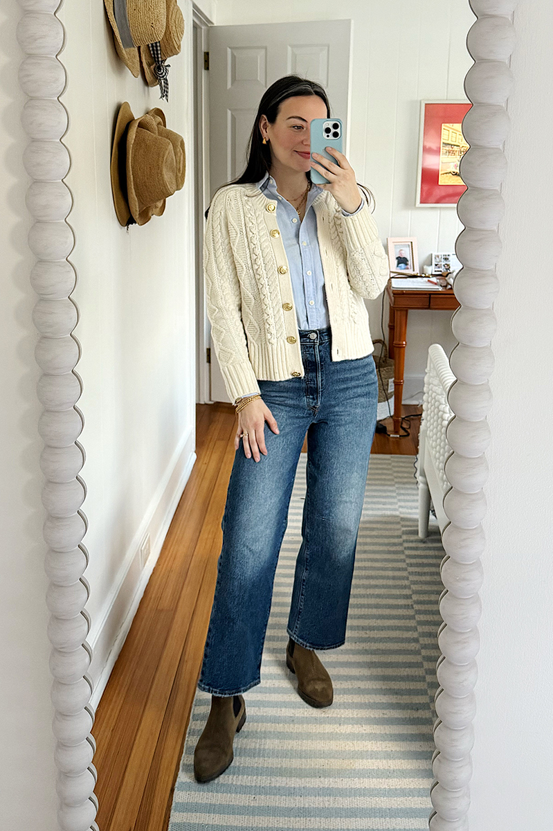 Carly Riordan taking a mirror selfie wearing a cream sweater, button down, and wide legged jeans.