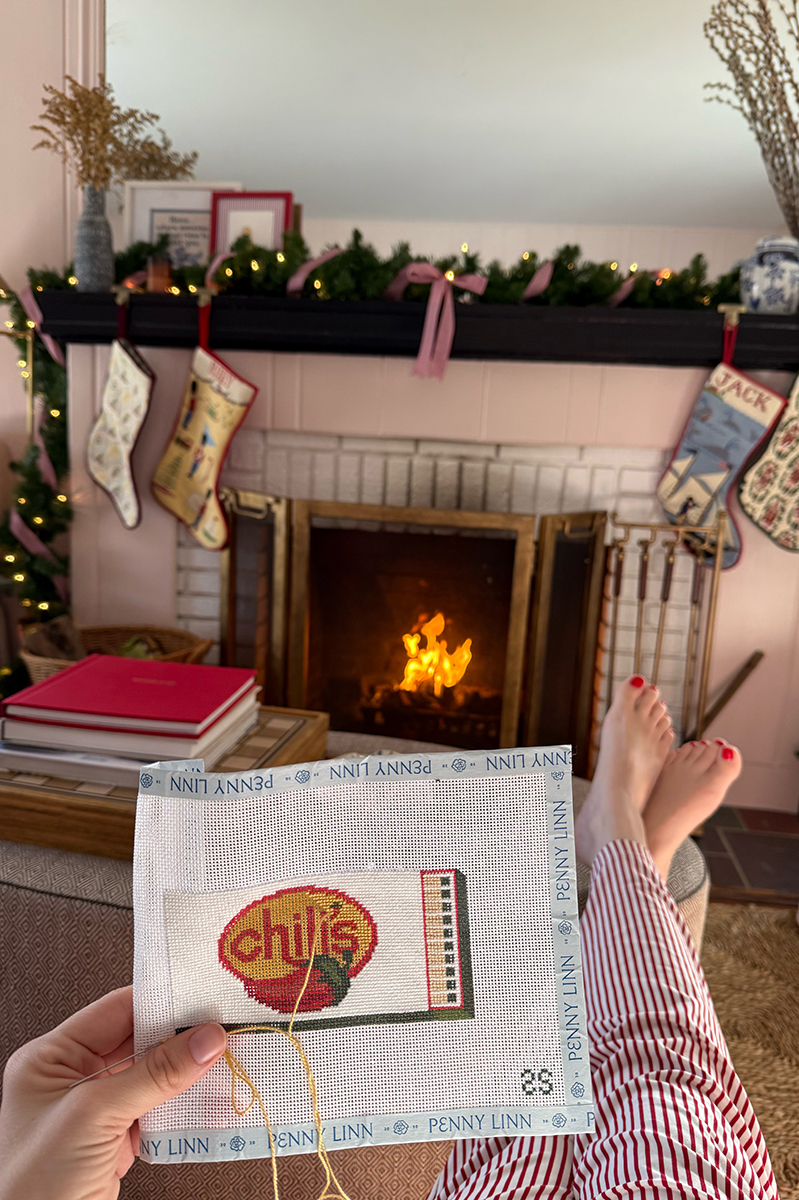 Carly needlepointing a Chili's logo canvas in front of a fire at Christmas